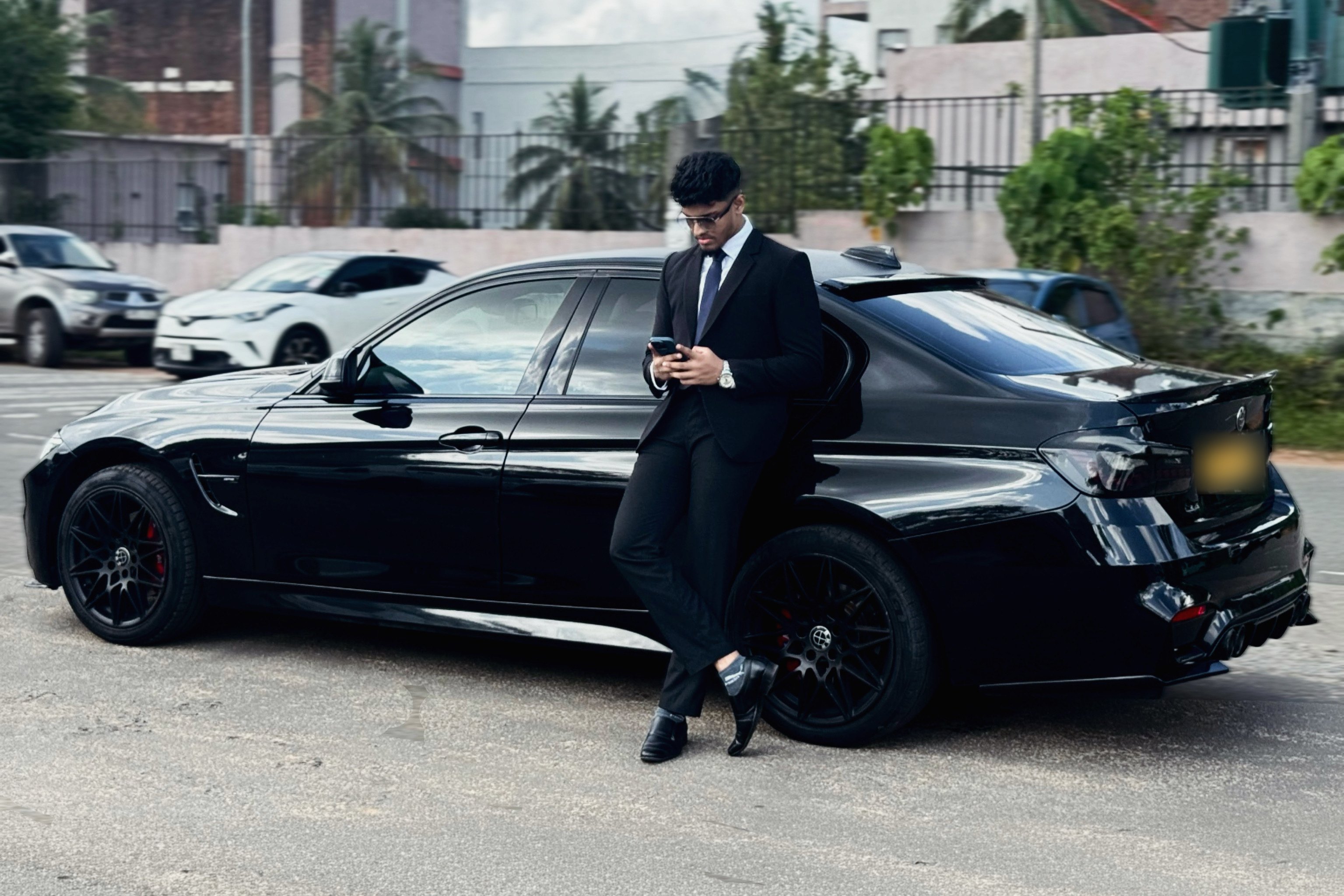 Lakindu Pahesara in a black suit leaning on a black BMW saloon, on a bright afternoon